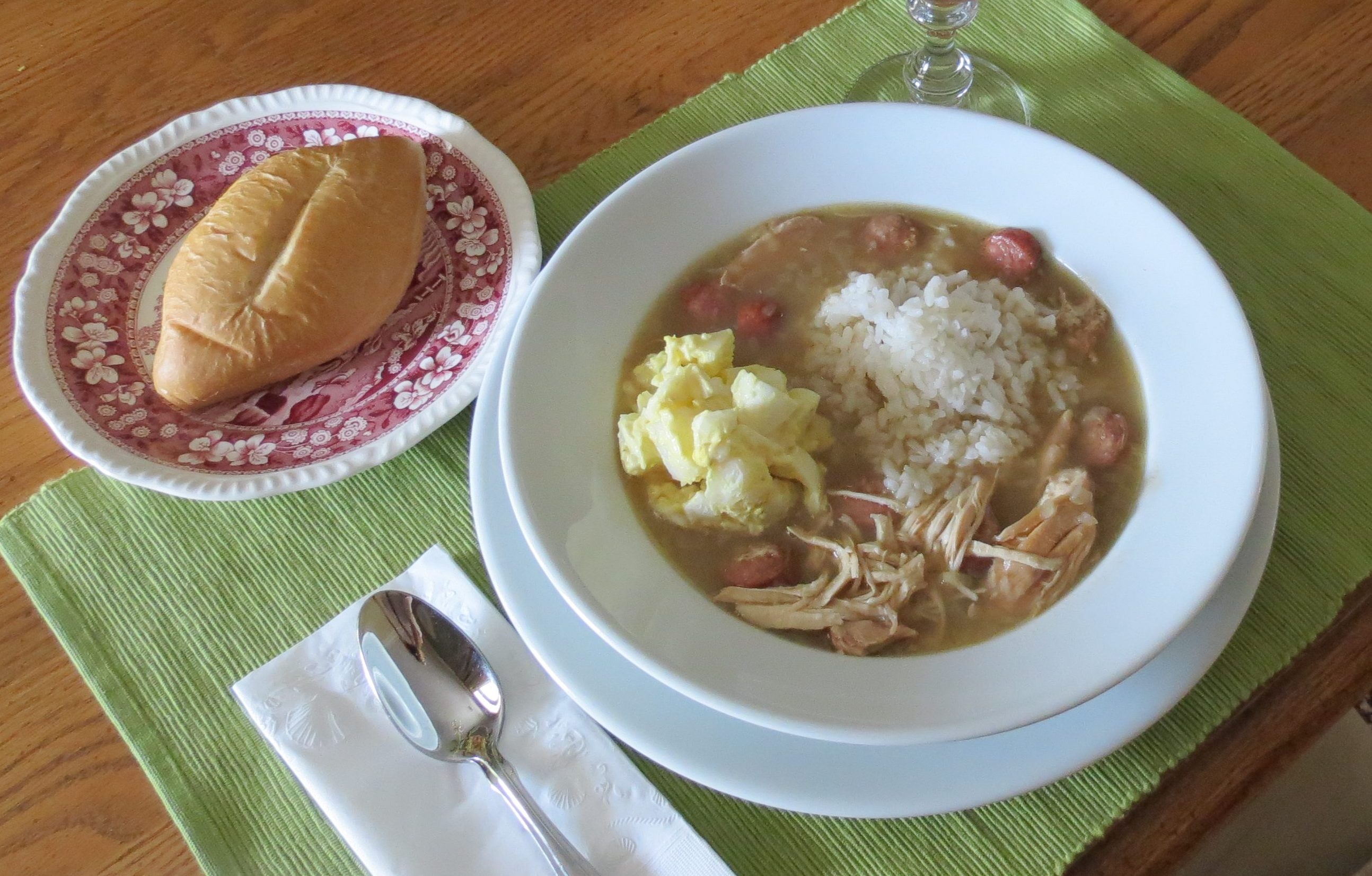 A white bowl of gumbo on a green place mat with a roll on a red and white plate.