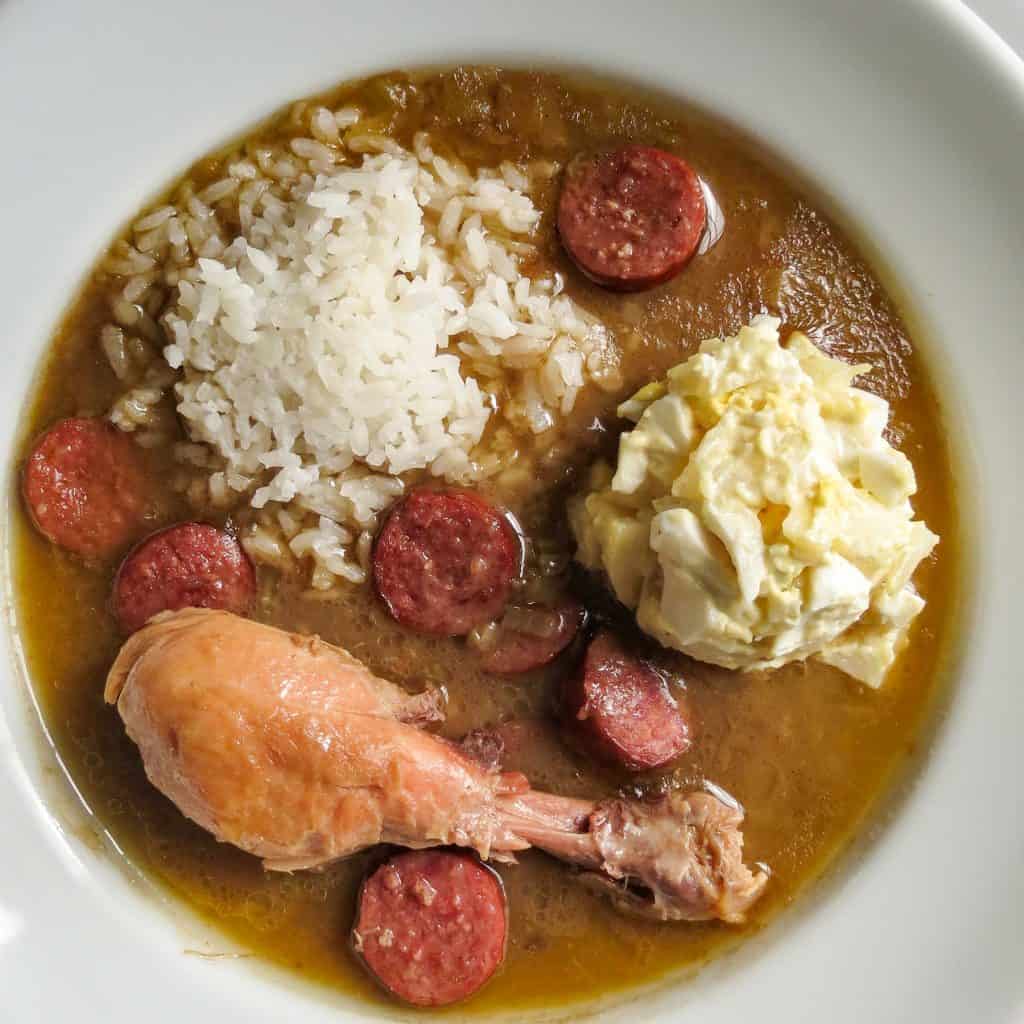 A white bowl full of rice, potato salad, sausage, and a chicken leg made from the Chicken Gumbo, Simply Classic Cajun recipe.