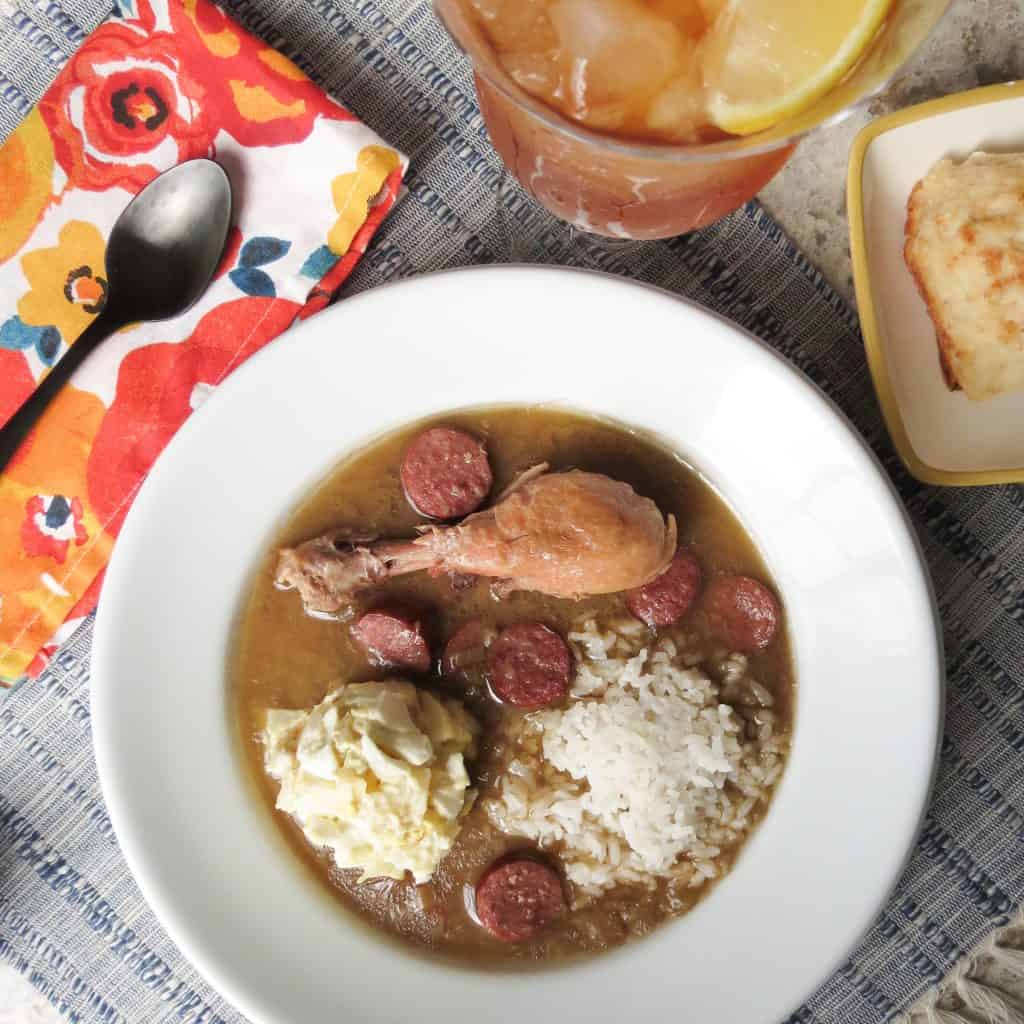 A white bowl full of rice, potato salad, sausage, and a chicken leg made from the Chicken Gumbo, Simply Classic Cajun recipe.