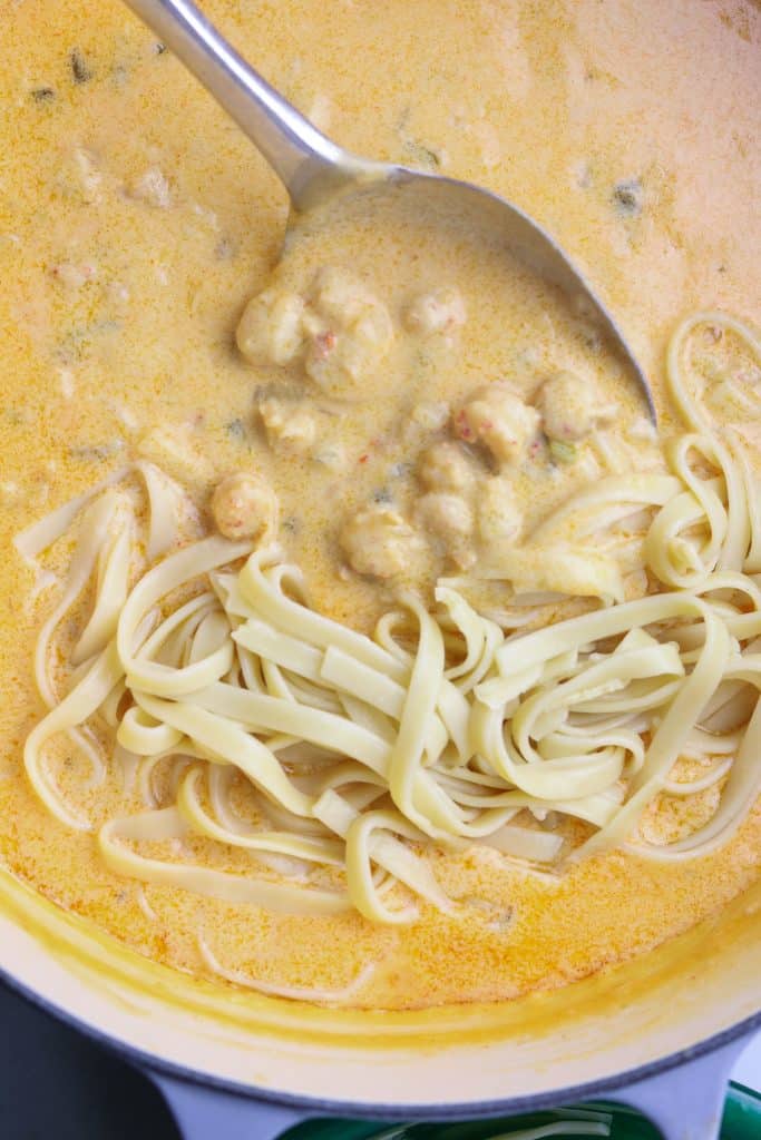 A spoon stirring in a pot of pasta, cream sauce, and crawfish.