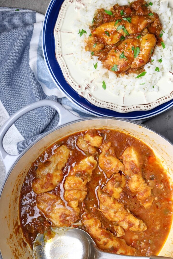 A plate of white rice topped with Fish In Red Gravy with a pot of fish cooked in a red sauce next to it.