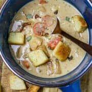 A blue bowl of Creamed Potato And Carrot Soup with a spoon on a wooden board with croutons.