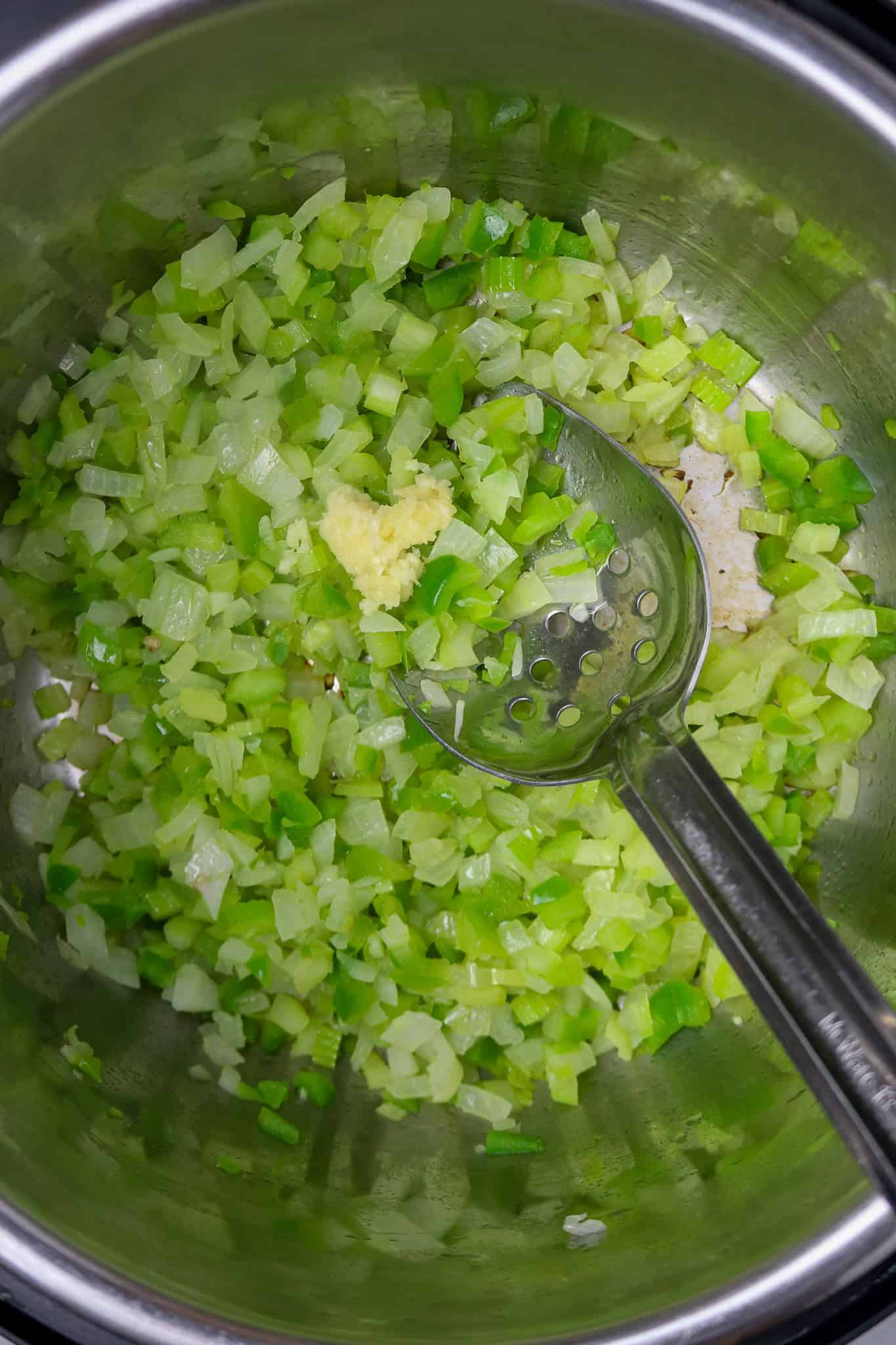 A long-handled silver spoon stirring onions, green pepper, and celery with garlic in a pot.