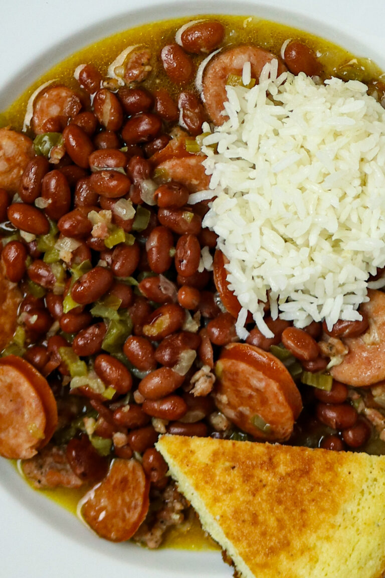 A plate of red beans with sausage and rice with a scoop of white rice and a wedge of corn bread on a white plate.