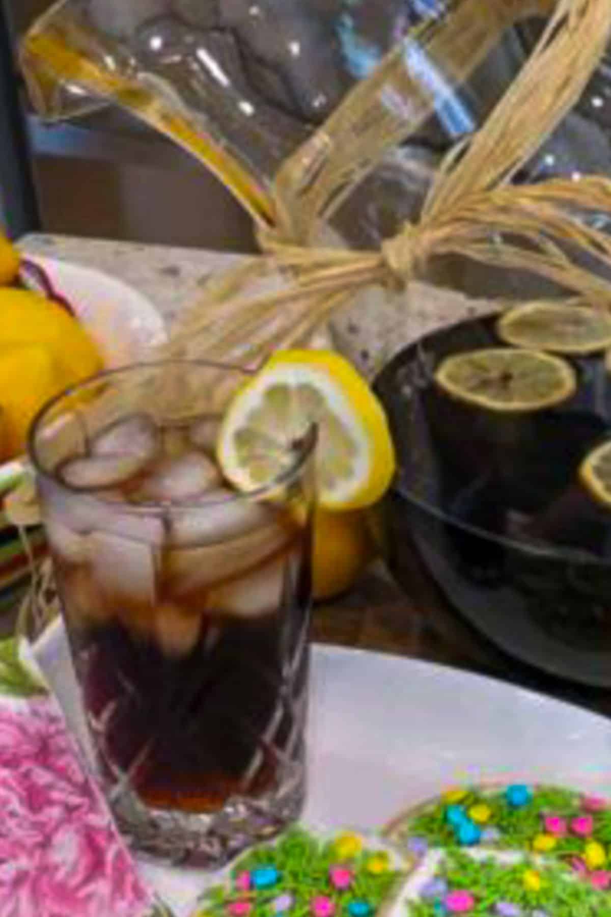 A clear glass of a dark liquid an ice with a lemon slice on the rim next to. a pitcher of root beer.
