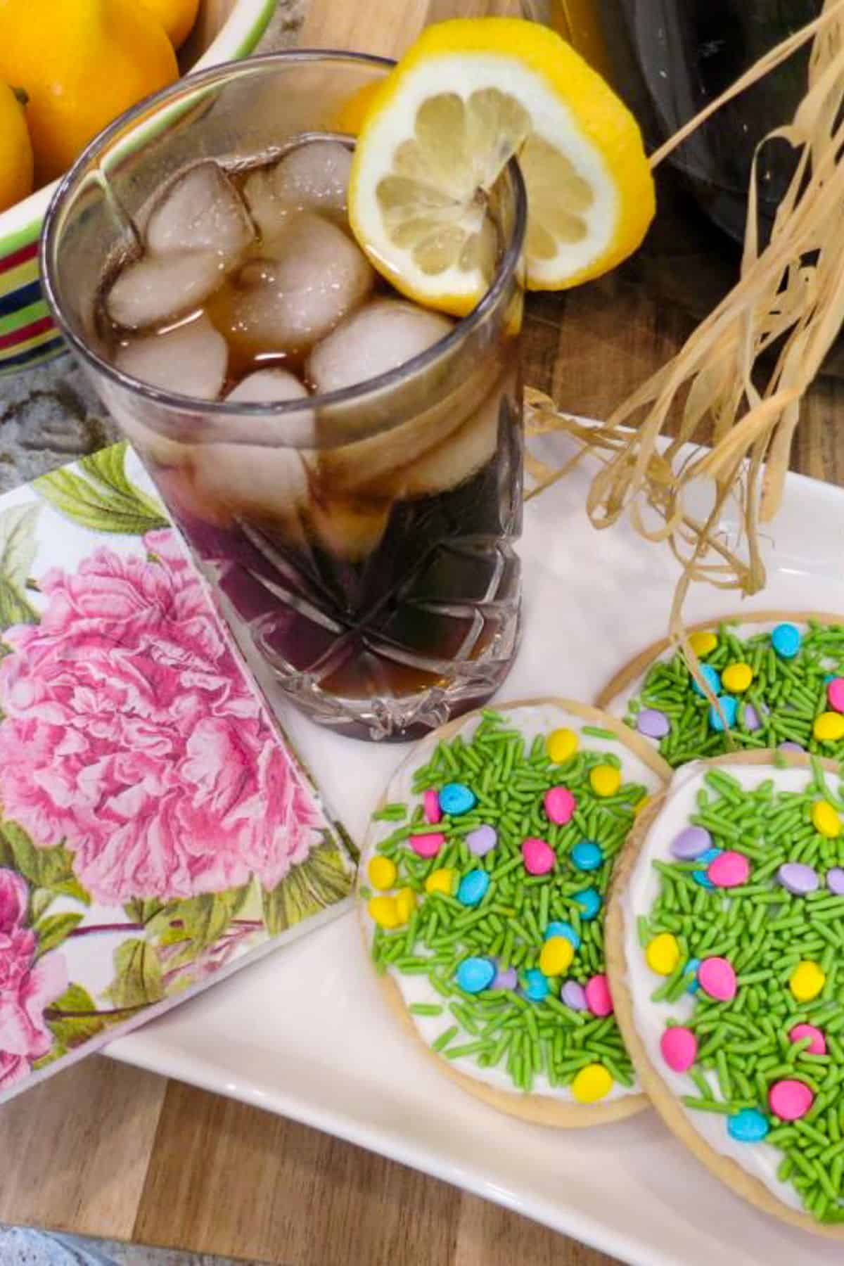 A clear glass of a dark liquid an ice with a lemon slice on the rim. next to Easter egg cookies.
