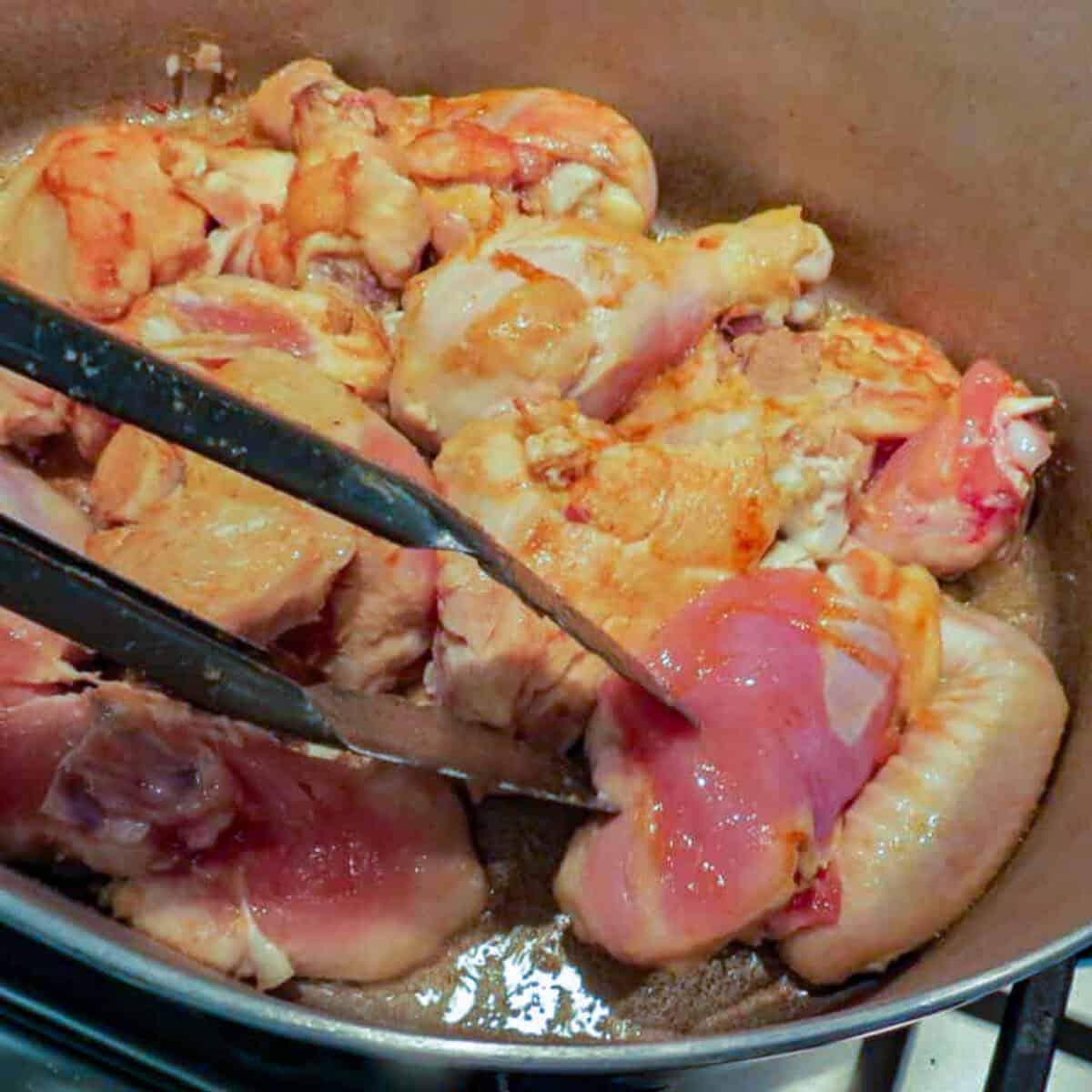 A pan with chicken in it and a pair of tongs to turn it.