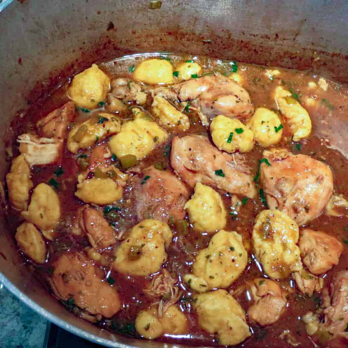 A large pot filled with chicken and dumplings and a dark gravy on a stove.