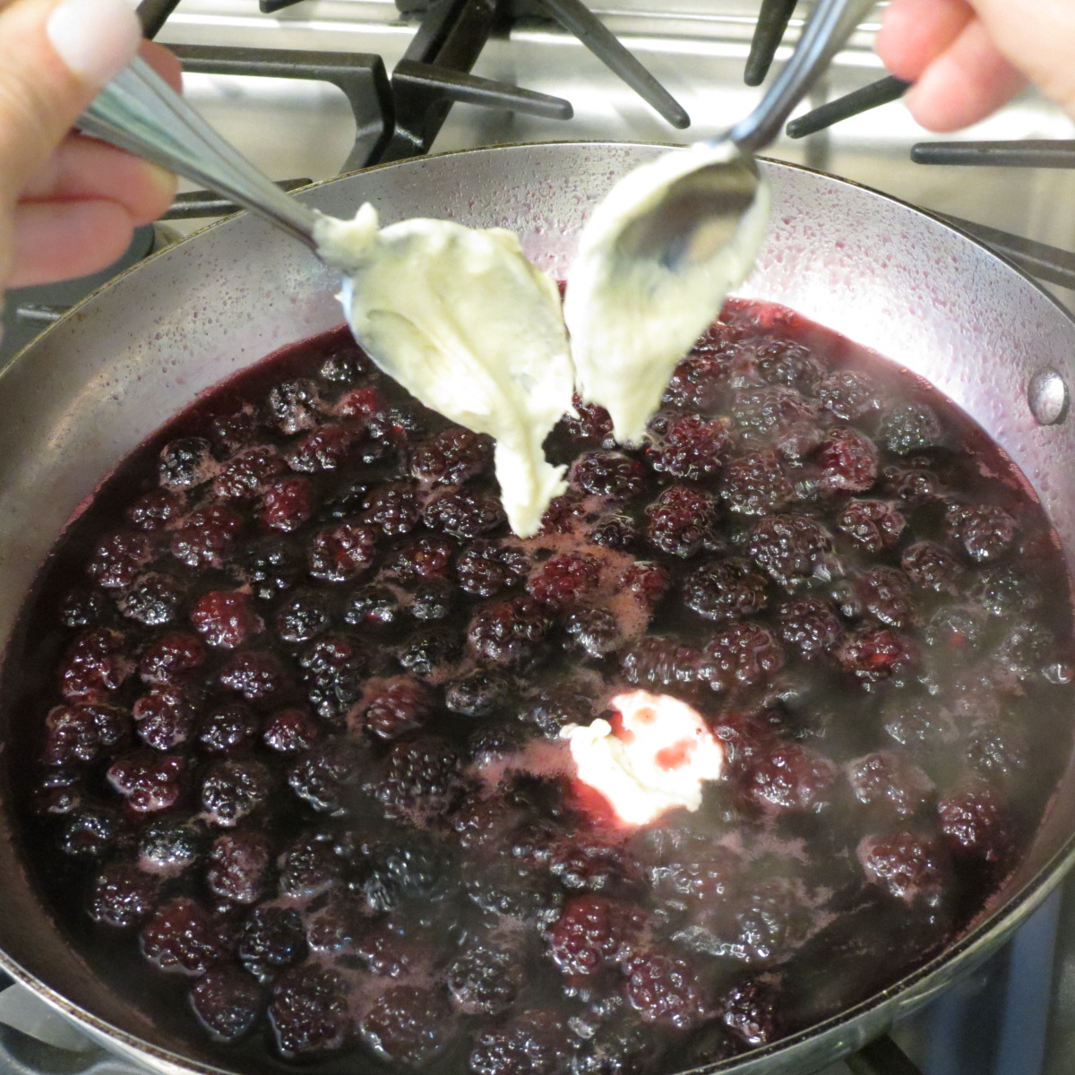 A saucepan of blackberry dumplings with 2 teaspoons dropping raw dough into the sauce.