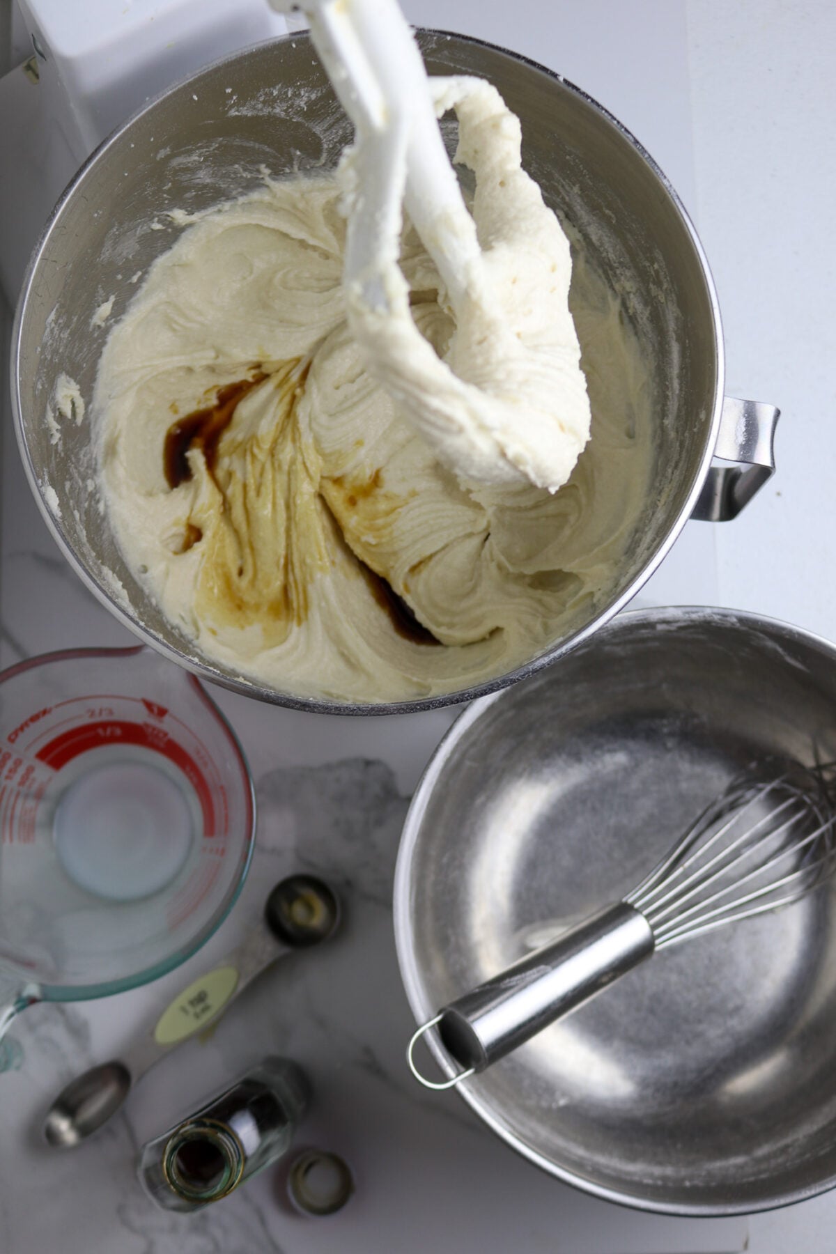 A silver bowl with cream and vanilla with beaters over it next to a silver bowl with and glass cup of cream.