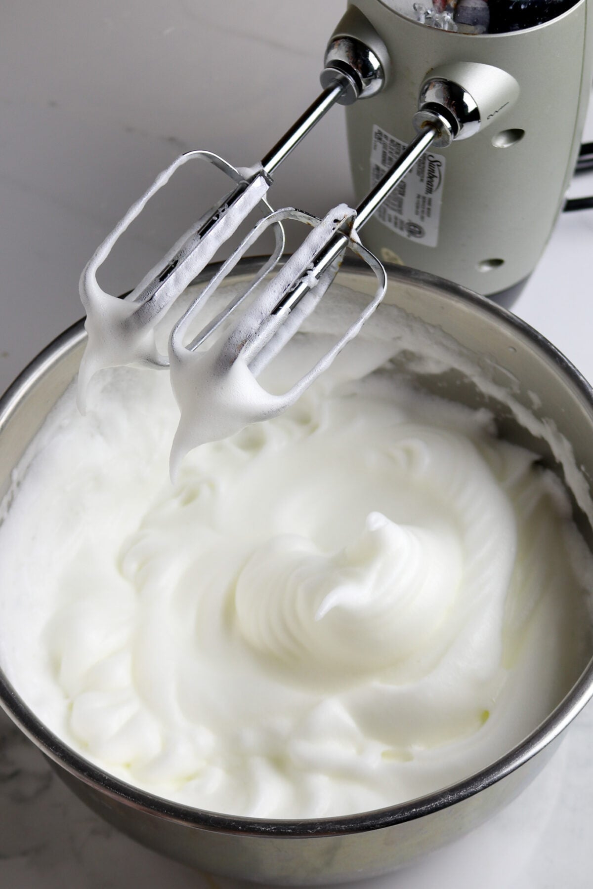 Two beaters over a silver bowl with whipped stiff egg whites.
