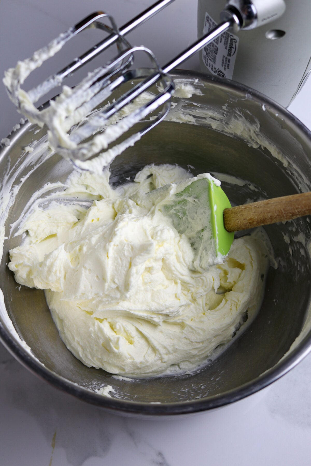 A silver bowl with a green spatula stirring mascarpone cheese powdered sugar, and vanilla.