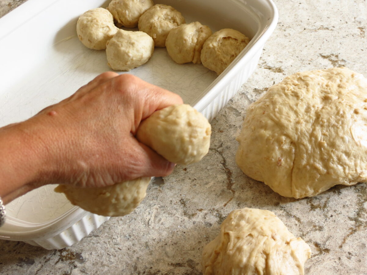 A hand squeezing dough into rolls over a pan of unbaked rolls.