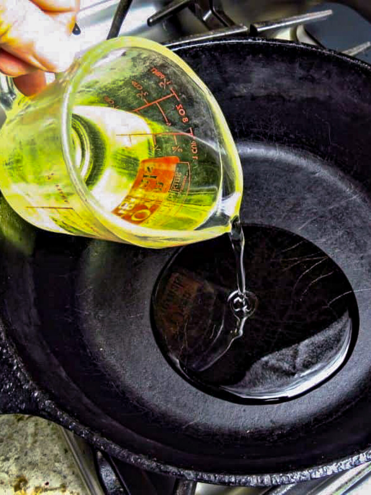 A cup of oil being poured into a black pot.