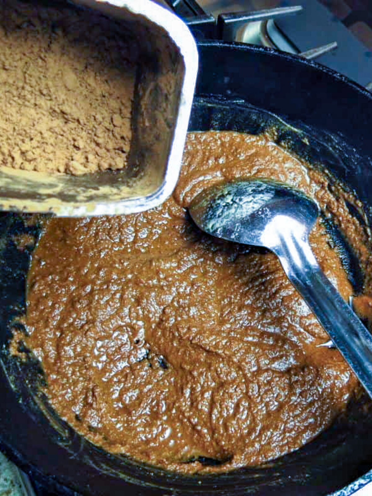 A black pot of medium-colored roux with a silver spoon and a container of cooa powder near near the top of the photo for a Cajun roux recipe.