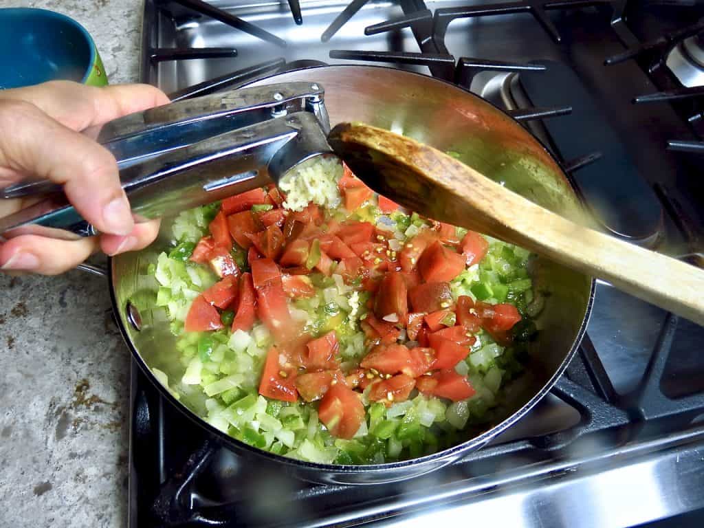 Adding fresh garlic from a press to cooking tomatoes, onions, and bell pepper in a pan.