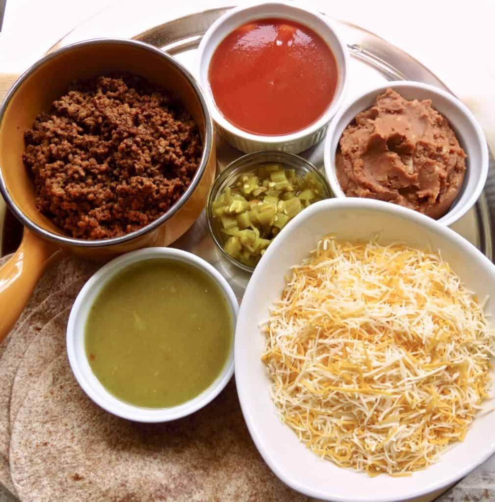 Ingredients of cheese, beef, refried beans, green chiles, and sauces for Easy Beef Enchiladas.