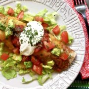 Easy Beef Enchilada resting on a bed of lettuce topped with sour cream, diced tomatoes, and avocados on a light gray plate.