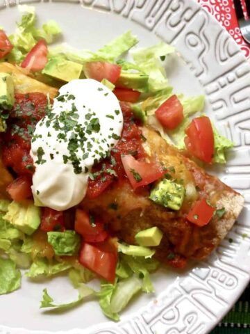 Easy Beef Enchilada resting on a bed of lettuce topped with sour cream, diced tomatoes, and avocados on a light gray plate.