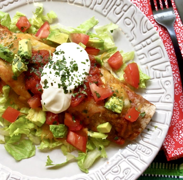 Easy Beef Enchilada resting on a bed of lettuce topped with sour cream, diced tomatoes, and avocados on a light gray plate.