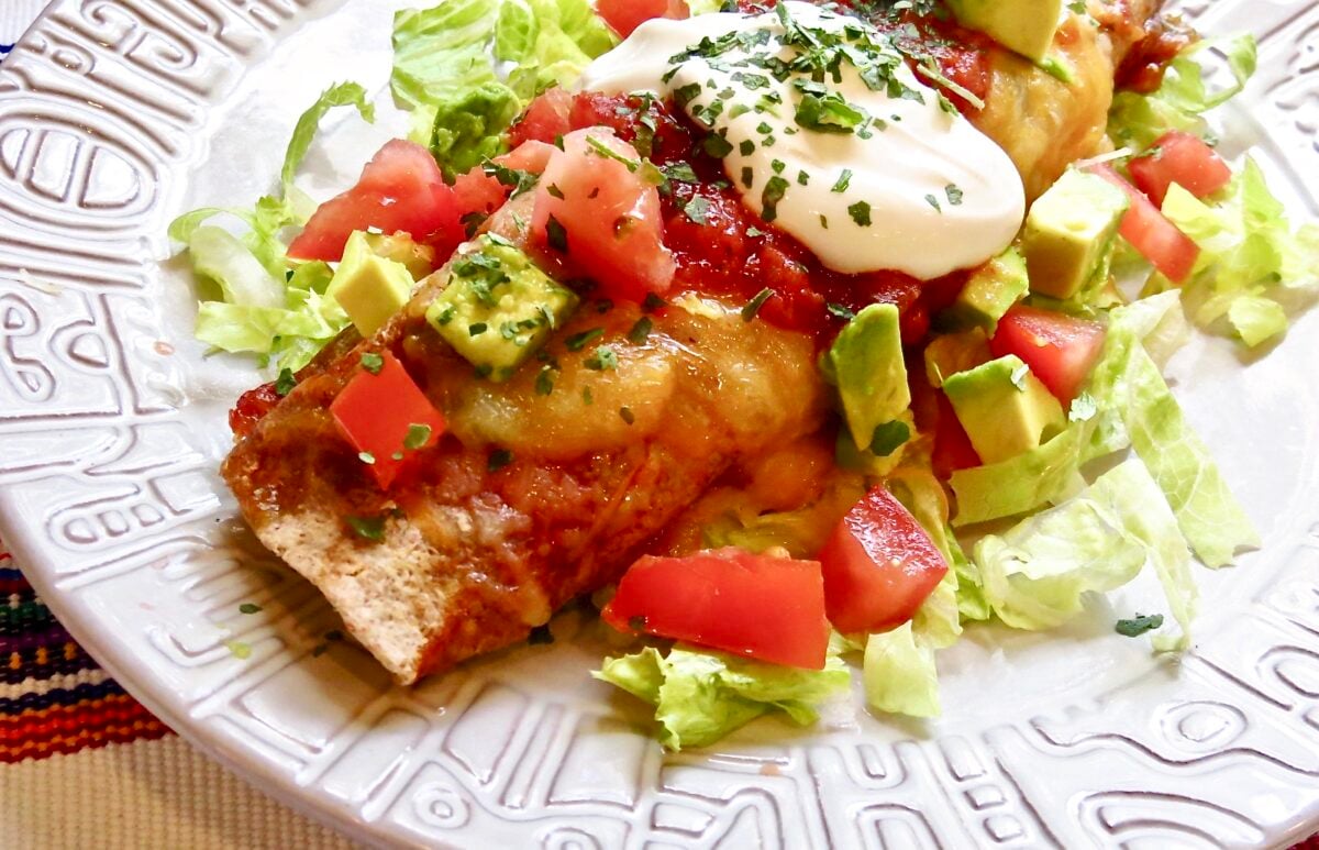 Easy Beef Enchilada resting on a bed of lettuce topped with sour cream, diced tomatoes, and avocados on a light gray plate.