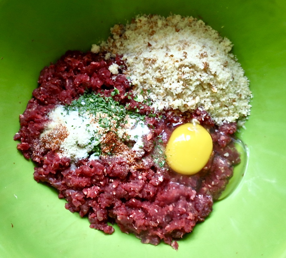 Ground meat with raw egg, seasonings, and breadcrumbs in green bowl.