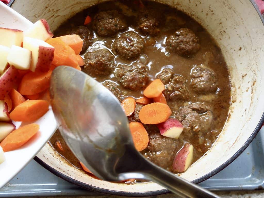 Sliced carrots being poured into a pot of meatballs and gravy.