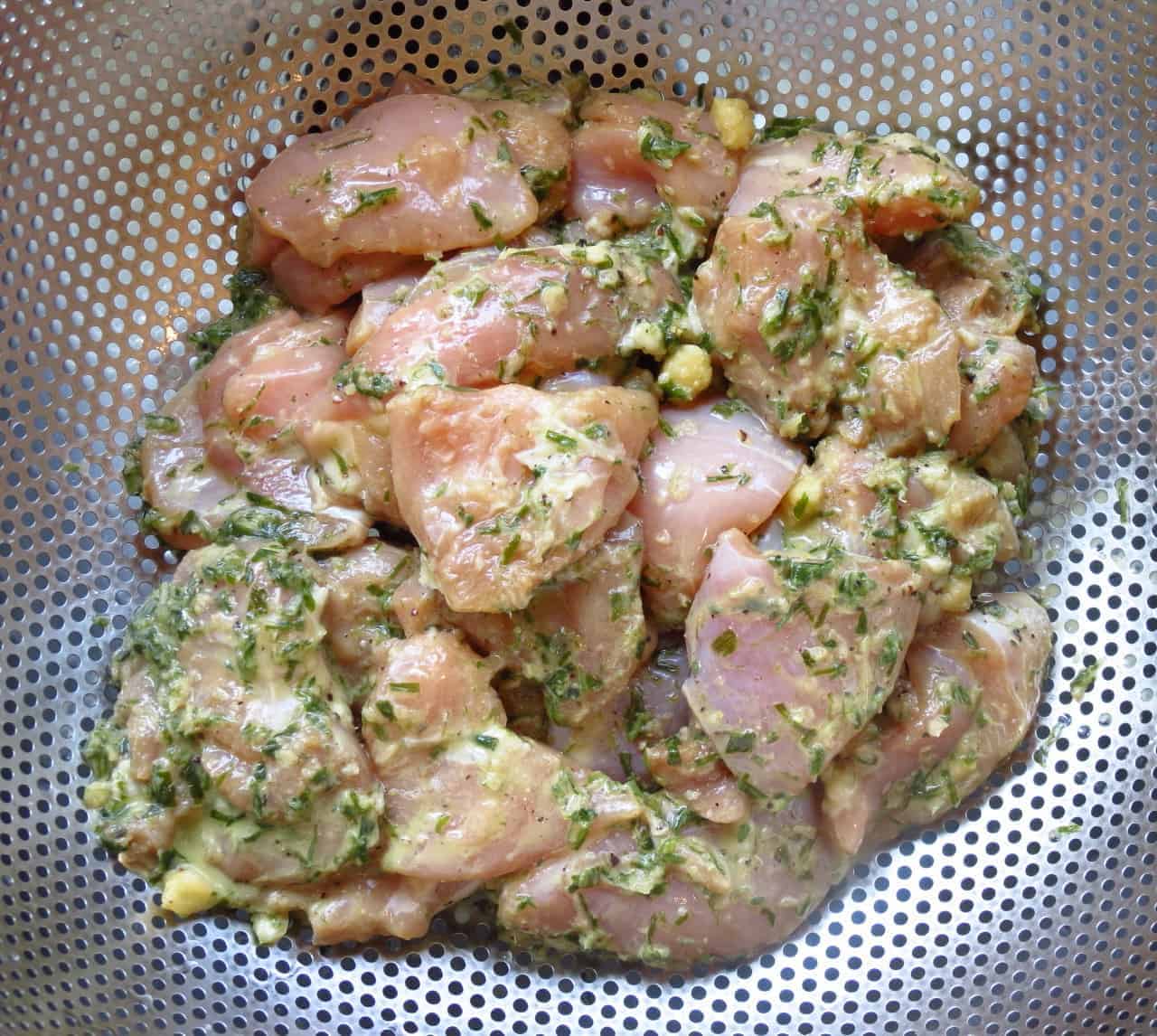 Marinated chicken thigh pieces in a stainless strainer.