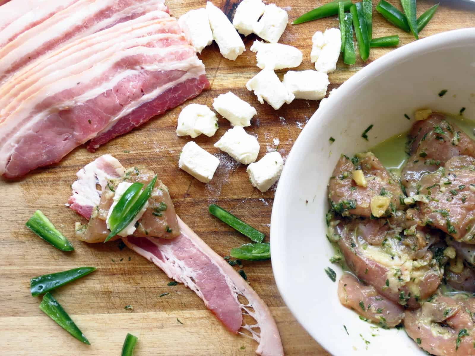 Slices of bacon with cubed cream cheese bits, sliced jalapeno peppers, and a bowl of marinated chicken cubes on a wooden board.