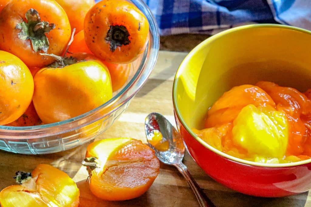 Persimmons in a bowl with a cut one on the side and a bow of peeled ones.