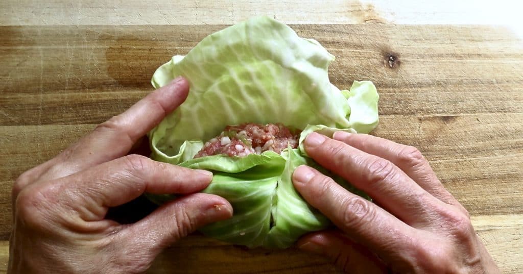 Two hands rolling up a roll of ground meat in a green leaf.