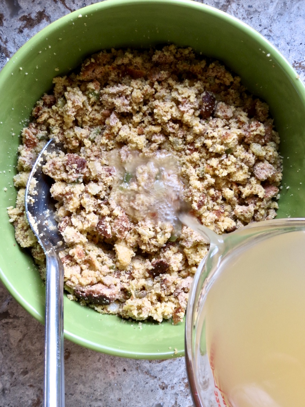 Chicken broth being poured into cornbread dressing mixture in a green bowl.