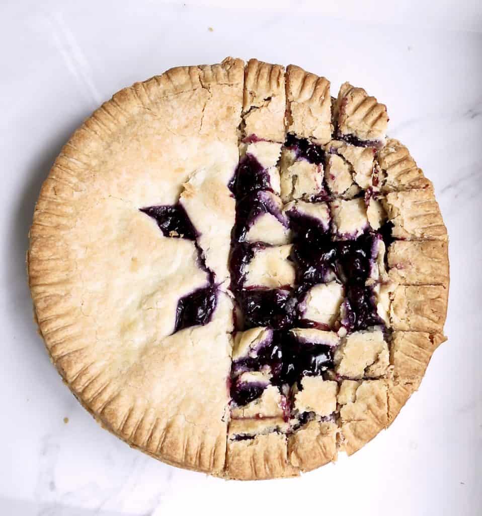 A blueberry pie on a white tray with half of it cut into small squares.