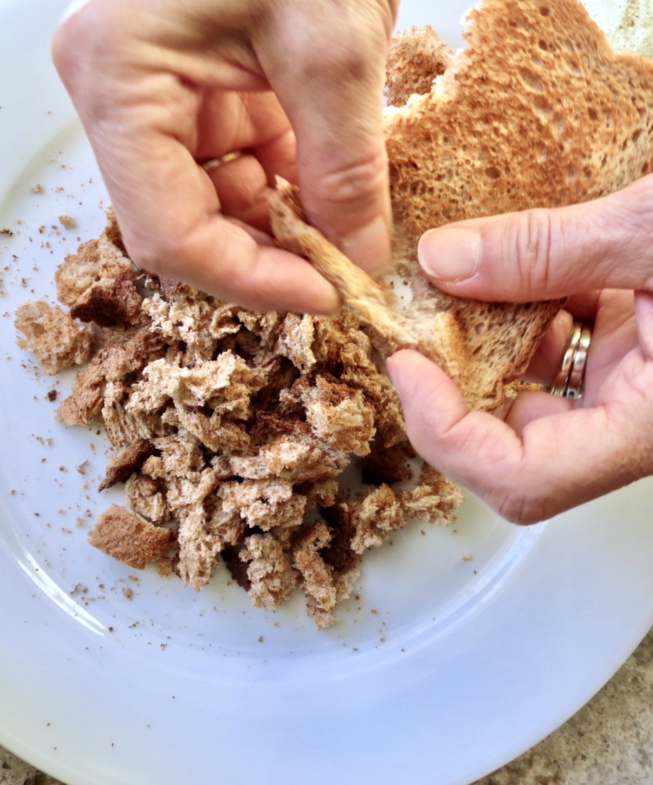 Two hands breaking up whole wheat toast to make bread crumbs for cornbread dressing.