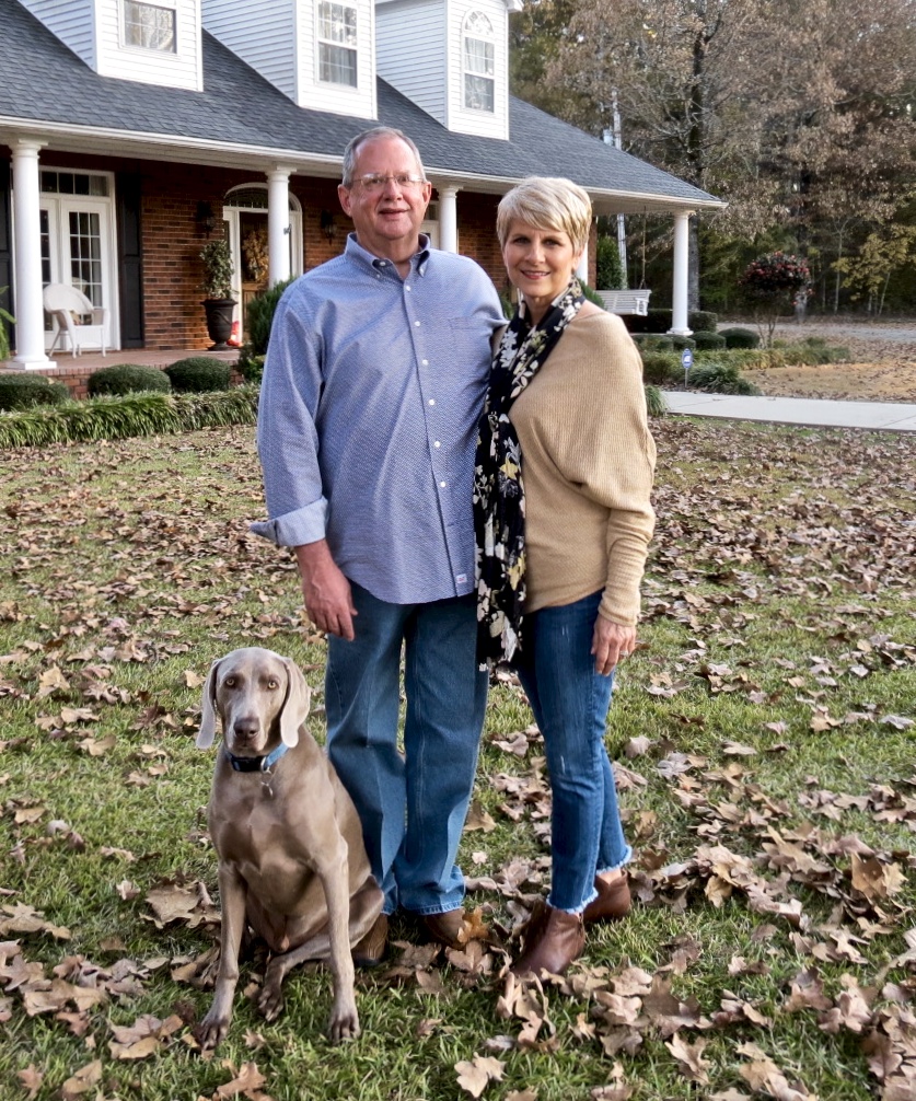 Picture of Steve and Kay standing in front of their brick home with their Weimaraner dog, Cord.