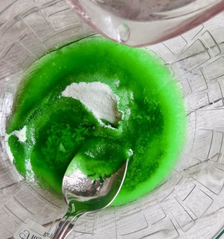A glass bowl of lime jello and water stirred by a spoon for Congealed Salad, Cherry Or Lime Flavored.