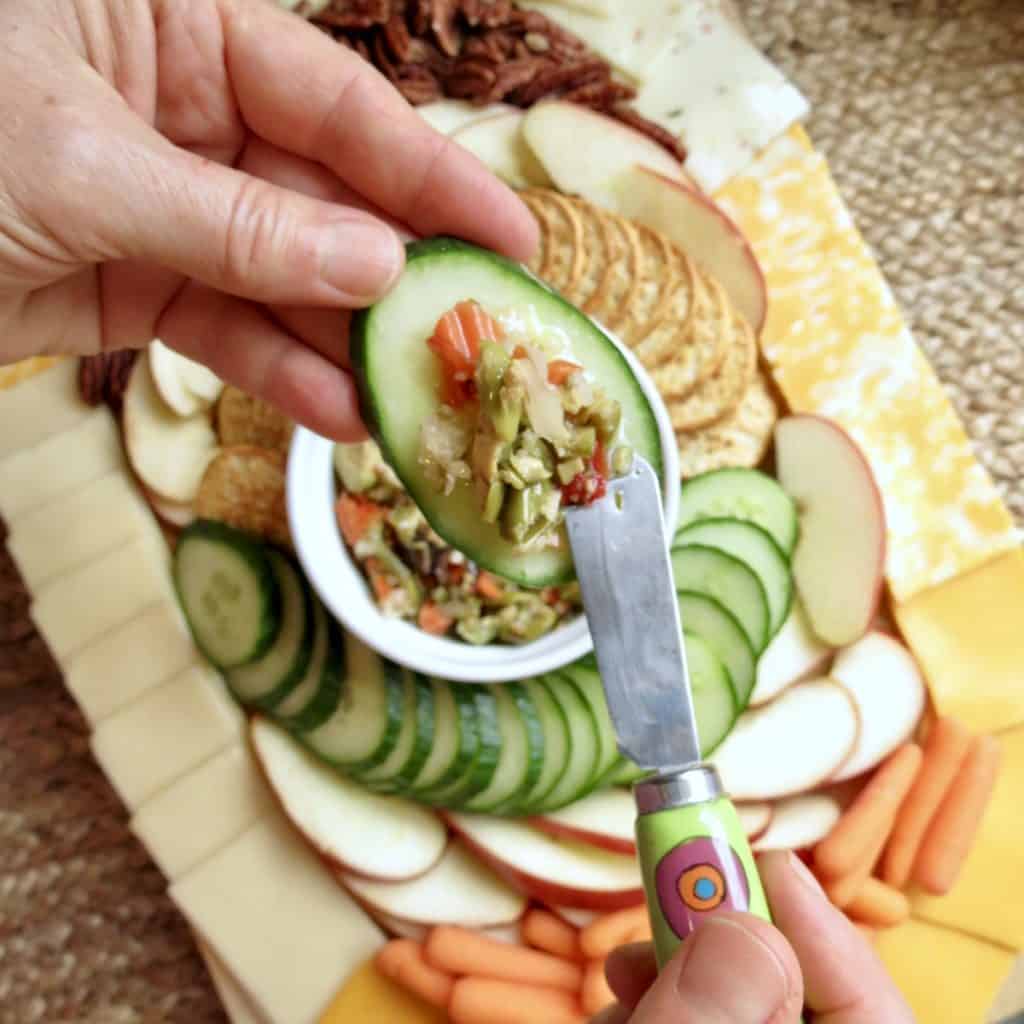 Sliced apples, pecans, crackers, sliced cucumber, baby carrots, and cheeses arranged in an oval around a bowl of olive salad on a cutting board for Easy Appetizers To Tease And Appease The Appetite.