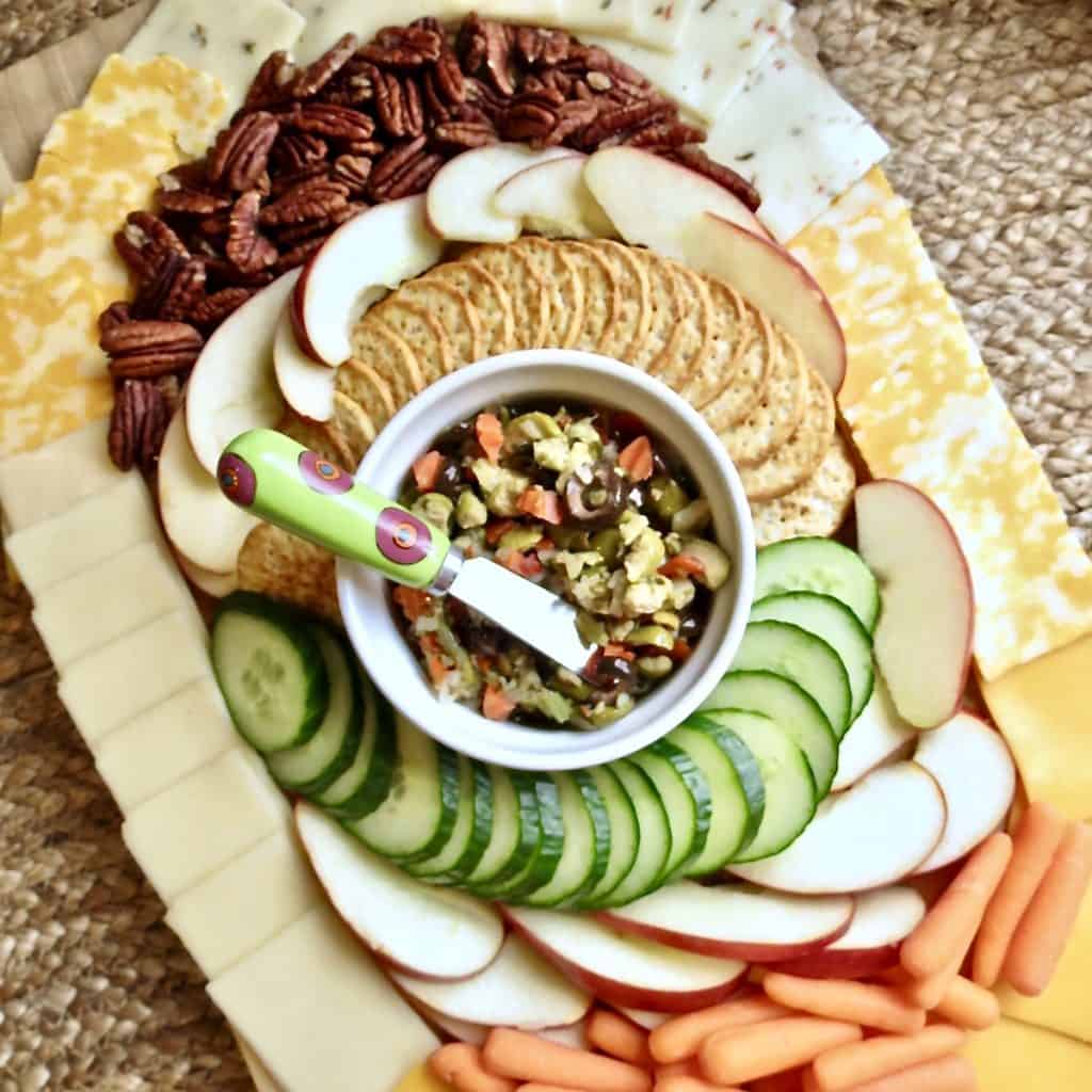 Sliced apples, pecans, crackers, sliced cucumber, baby carrots, and cheeses arranged in an oval around a bowl of olive salad on a cutting board for Easy Appetizers To Tease And Appease The Appetite.
