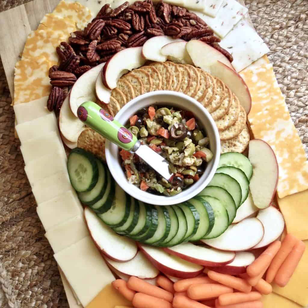 Sliced apples, pecans, crackers, sliced cucumber, baby carrots, and cheeses arranged in an oval around a bowl of olive salad on a cutting board for Easy Appetizers To Tease And Appease The Appetite.