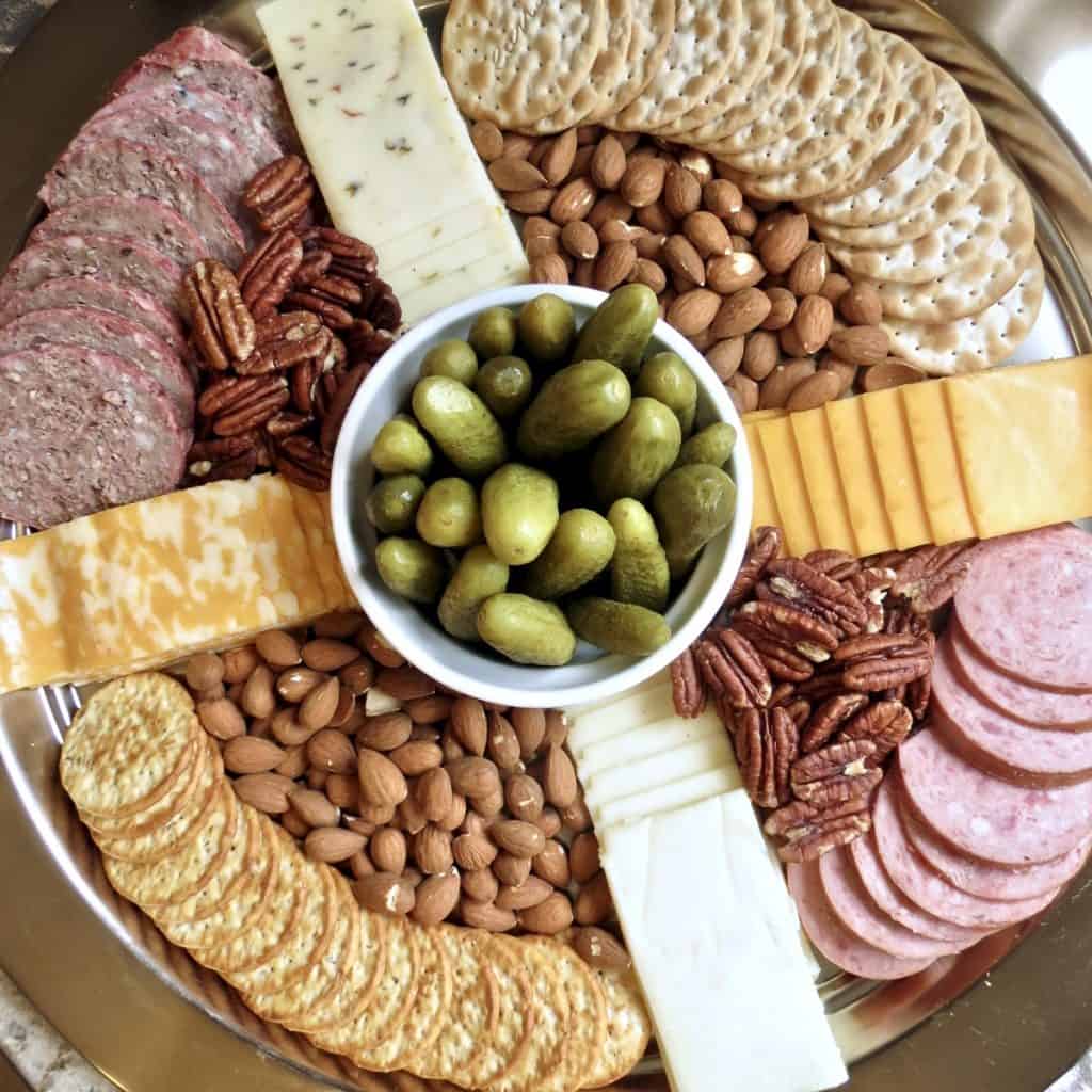 Sliced salami, summer sausage, nuts, sliced cheese, crackers arranged around a dish of pickles on a tray for Easy Appetizers To Tease And Appease The Appetite.