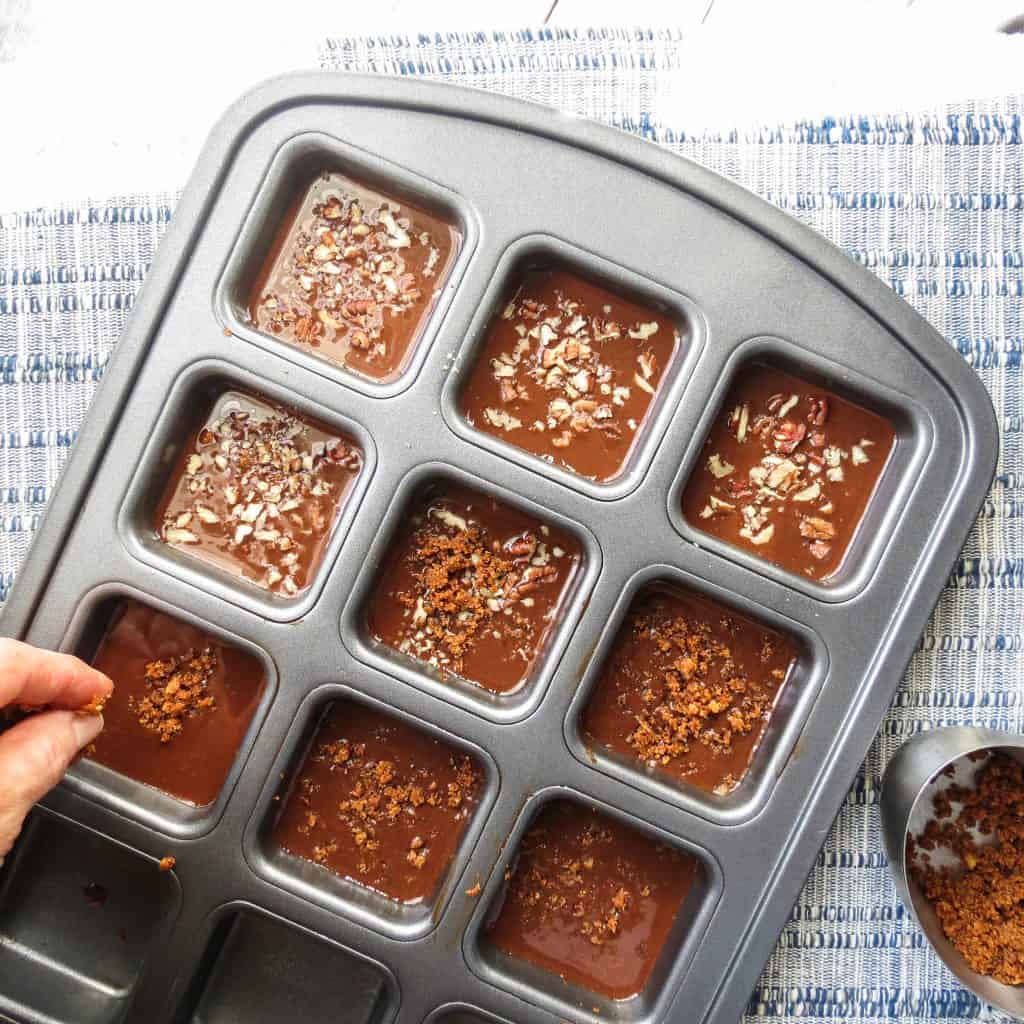 A hand sprinkling Cajun cracklins and pecans onto Fat Bombs in square molds.