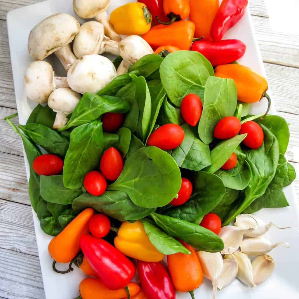 Fresh spinach, mushrooms, colored mini peppers, and grape tomatoes on a white tray.