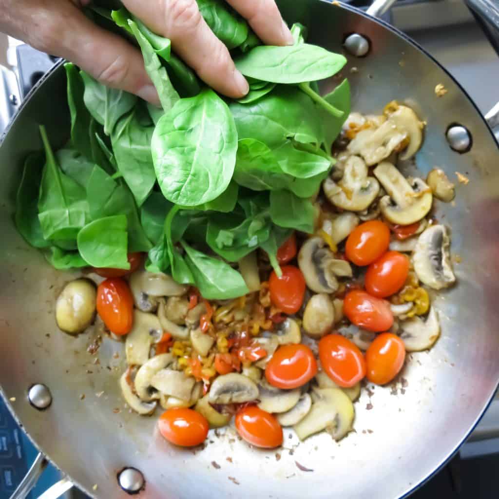 A handful of fresh green spinach thrown into a pan of sautéed peppers, mushrooms, and grape tomatoes with a wooden spoon for Spinach Medley, A Healthy Spinach Side Dish.