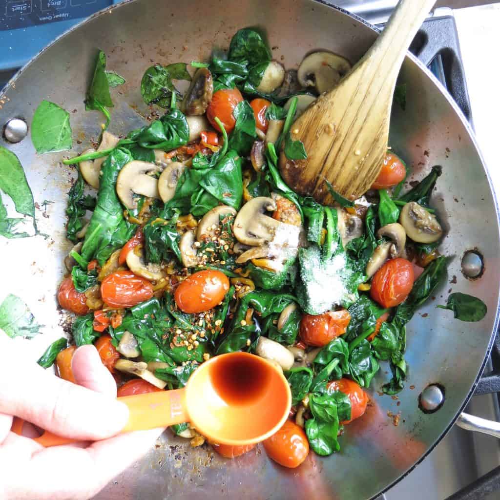 A Pan of sautéed spinach, peppers, mushrooms, and grape tomatoes with a wooden spoon for Spinach Medley, A Healthy Spinach Side Dish.