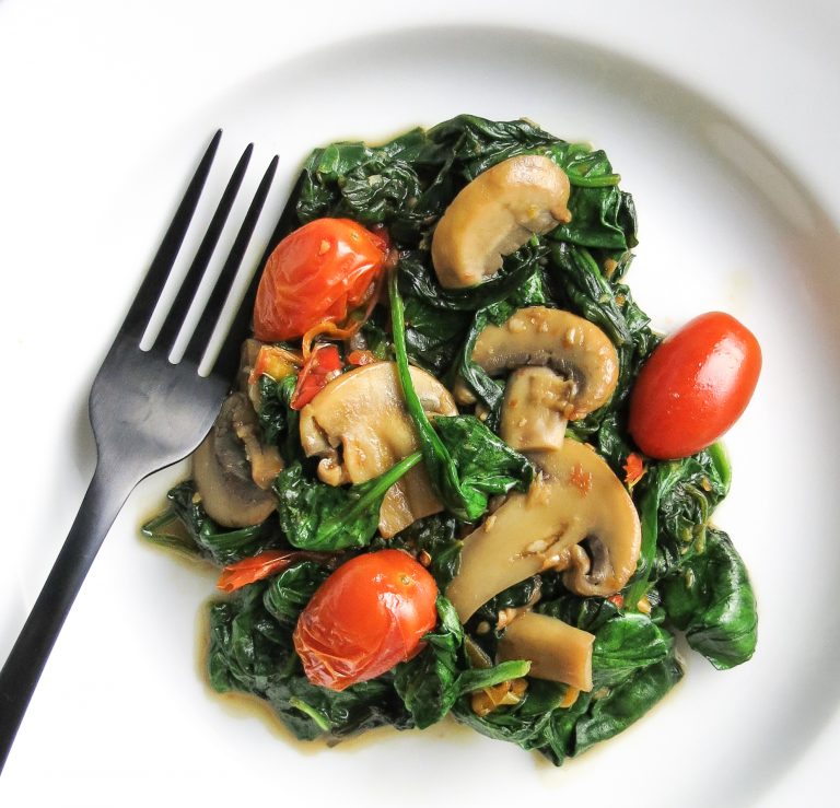A white bowl with a black fork holds a serving of sautéed spinach, Mushrooms, grape tomatoes, and sweet peppers for a Spinach Medley, A Healthy Spinach Side Dish.