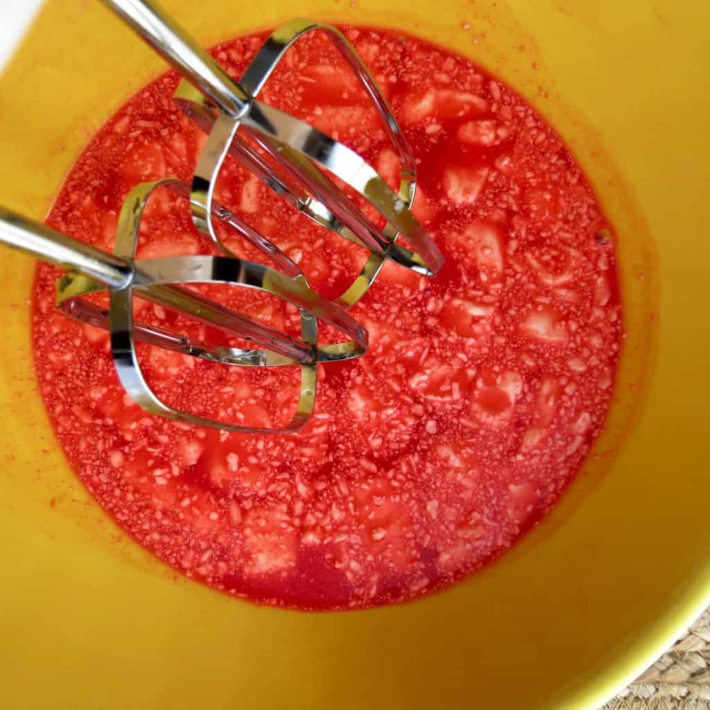 Electric beaters in a yellow bowl of strawberry jello and cream cheese for Keto Strawberry Jello Dessert.