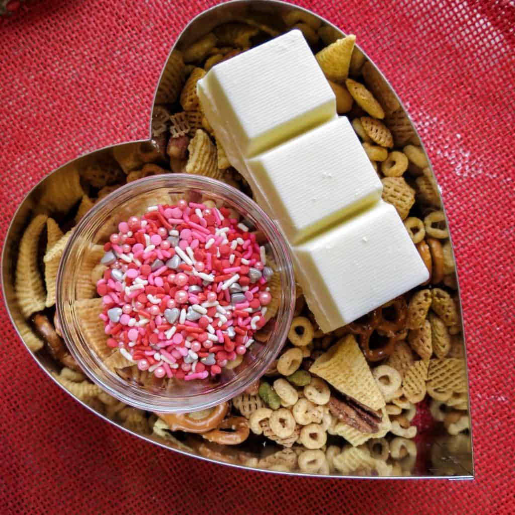 Ingredients of cereal, sprinkles, and white chocolate in a heart shape mold on a red placemat.