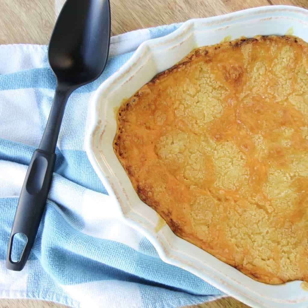 A casserole filled with Amy's Corn Casserole that has a browned topping of cheese with a black serving spoon on a blue and white towel.