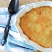 A casserole filled with Corn Casserole that has a browned topping of cheese with a black serving spoon on a blue and white towel.
