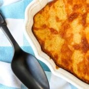 A casserole filled with Corn Casserole that has a browned topping of cheese with a black serving spoon on a blue and white towel.